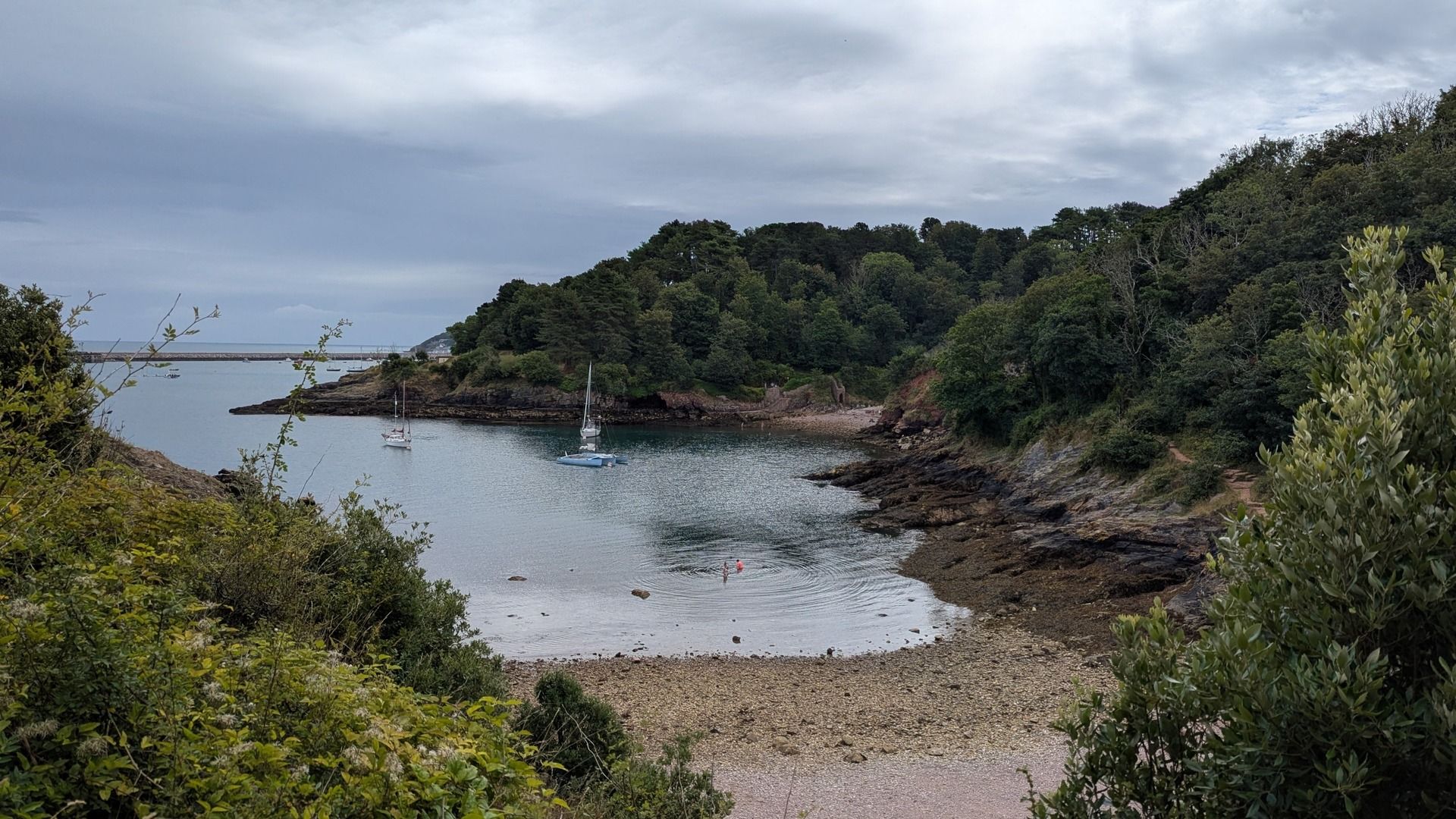 A picture taken from an elevated position looking down at a peaceful cove. Two people are wading in the water and three boats are anchored just off the shore